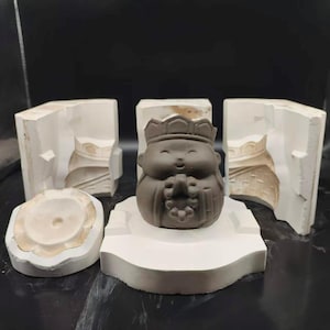 May include: A dark brown ceramic Buddha figurine with a crown, displayed with white plaster moulds. The Buddha sits on a white base. The moulds are in different shapes, indicating a casting process, likely for ceramics.