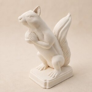 May include: A matte off-white squirrel figurine standing on a rectangular base. The squirrel is depicted in an upright position, holding a round nut between its front paws, with its bushy tail curved upward behind its back and detailed fur texture.