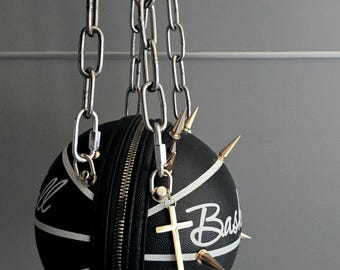 May include: A spherical black handbag designed like a basketball with white lines and the text "Basketball". The bag features gold-toned metal spikes, a central zipper closure, and a silver-toned metal chain strap with a small cross charm.