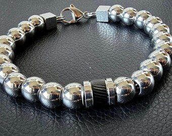 May include: A bracelet composed of polished silver-toned spherical beads, featuring a central black cylindrical bead with vertical grooves, two square silver-toned spacers, and a lobster claw clasp closure mechanism.
