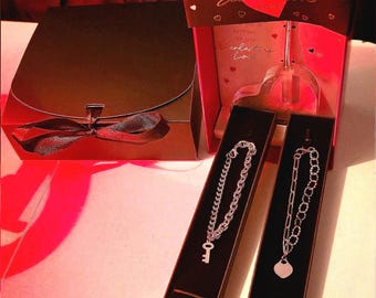 May include: A red artificial rose in a rectangular box labeled Eternal Love, a black gift box with a ribbon, two silver-toned chain bracelets with key and heart charms in narrow boxes, and a card labeled Astrious Studio with text.