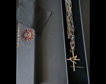 May include: A silver-toned chain necklace featuring a gold-toned pendant shaped like a bird integrated into a cross. The necklace is presented in a long black rectangular box alongside a black polishing cloth. The box lid displays the text ASTRIUS STUDIO.