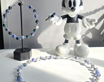May include: A beaded bracelet and necklace set with blue, white, and green stones. The bracelet is displayed on a black stand. A Mickey Mouse figurine is in the background, holding a matching beaded ring. The jewelry is wire-wrapped.