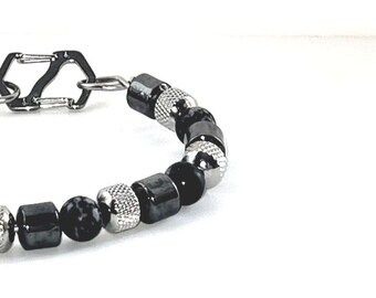May include: A close-up of a bracelet featuring a mix of black, silver, and patterned beads. The bracelet has a silver clasp and a variety of bead shapes, including round, cylindrical, and textured. The beads are strung together to form a stylish accessory.