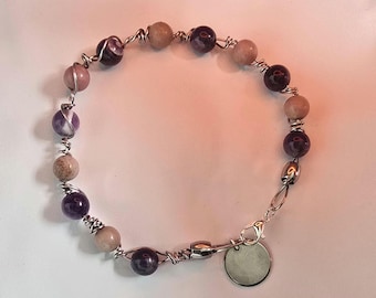 May include: A bracelet featuring alternating round purple amethyst beads and tan speckled stone beads, each individually wrapped in silver-toned wire. The piece includes a silver-toned lobster claw clasp and a small, round, blank silver-toned metal charm.