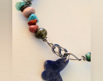 May include: A bracelet featuring a series of multi-colored, irregular chip-shaped stones in shades of blue, green, red, pink, yellow, and purple, strung on a wire. A dark blue butterfly-shaped stone charm hangs from a silver-toned metal clasp.