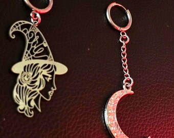 May include: A pair of silver-toned hoop earrings. One earring features a dangling cutout charm of a person wearing a pointed hat with intricate line details. The other earring has a chain link drop ending in a textured crescent moon charm.