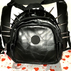 May include: Black backpack with silver spikes. The bag features a patchwork design and a small zipped front pocket. The straps and back of the backpack are also black.