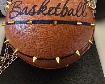 May include: A round, brown, textured basketball-shaped handbag featuring black lines, gold-tone metal spikes, and the word "Basketball" in black script. The bag includes a gold-tone chain strap with a brown leather-like shoulder pad attachment.