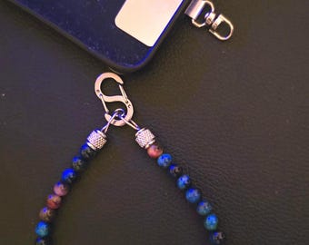 May include: A beaded wrist strap featuring small, round, multi-colored blue and purple stones interspersed with silver-toned metal beads. The strap is attached to a silver-toned carabiner clip, which connects to a black phone case.