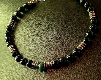 May include: A beaded necklace featuring smooth black spherical beads interspersed with metallic copper-toned ribbed cylindrical spacers. A single dark green bead is positioned near the bottom, and the necklace includes a silver-toned chain.