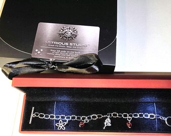 May include: A silver-toned chain bracelet with four charms, including a flower, a rose, and two red beads, presented in a rectangular red box with an LED light. A black card with the text ASTRIOUS STUDIO and a black ribbon are placed behind the box.