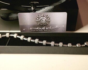 May include: A silver-toned metal bracelet featuring a series of cylindrical and textured beads, presented in a long black rectangular box. Behind the box sits a card with the text AETRIOUS STUDIO and the slogan Break the Static. Wear the Shield.