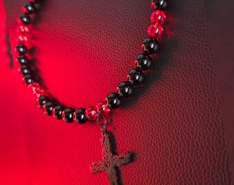 May include: A beaded necklace featuring alternating glossy black and translucent red spherical beads. A dark metal cross pendant with a textured, branch-like surface hangs from the center. The necklace includes a silver-toned metal clasp.