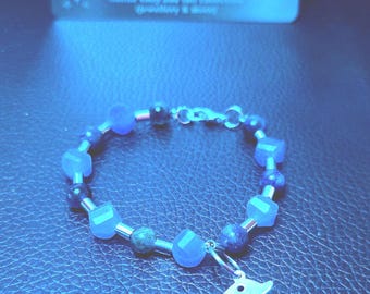 May include: A blue beaded bracelet featuring a silver-toned dolphin charm and alternating faceted and smooth round beads. In the background, a rectangular card displays the text Astrious Studio, Break the Static, Wear the Shield.
