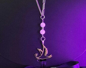May include: A silver-toned necklace featuring a mixed-link chain, two pale pink spherical beads, and a pendant shaped like a bird in flight holding a small branch. The necklace is displayed against a dark surface with a black ribbon.