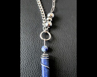 May include: A necklace featuring a silver-toned chain with a mix of curb and paperclip links. The pendant consists of a cylindrical blue lapis lazuli stone wrapped in silver-toned wire, topped with a small round lapis bead and three silver-toned spheres.