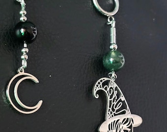 May include: A pair of mismatched silver-tone hoop earrings. One earring features a black bead and a silver crescent moon charm. The other earring features a green bead and a silver charm depicting a profile of a person wearing a witch hat.