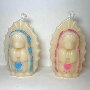 May include: Two ivory-colored candles shaped like a religious figure. One candle has blue accents and a blue heart, the other has pink accents and a pink heart. Both candles have a white wick.