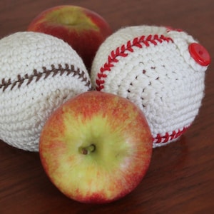 Puede incluir: Tres pelotas de béisbol de crochet, una con costuras rojas y un botón rojo, y dos con costuras marrones. Hay dos manzanas rojas en el fondo.