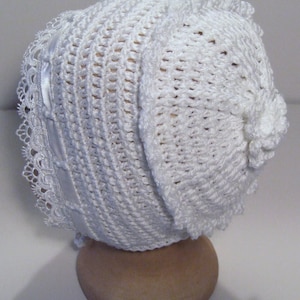 May include: White crocheted baby bonnet with a lace trim and a satin ribbon. The bonnet has a flower detail on the side.
