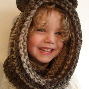 Puede incluir: Una bufanda de punto marrón y beige con capucha y orejas de oso. La bufanda la lleva puesta un niño con el pelo rubio.