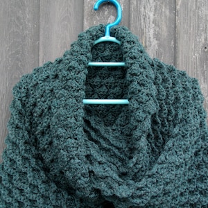 May include: A teal blue crocheted scarf hanging on a blue plastic hanger against a grey wooden background.