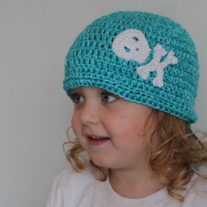 Puede incluir: Un niño pequeño con un gorro de punto de color azul turquesa con un diseño de calavera y huesos cruzados blancos. El gorro tiene una cola larga que cuelga por la parte posterior del cuello del niño.