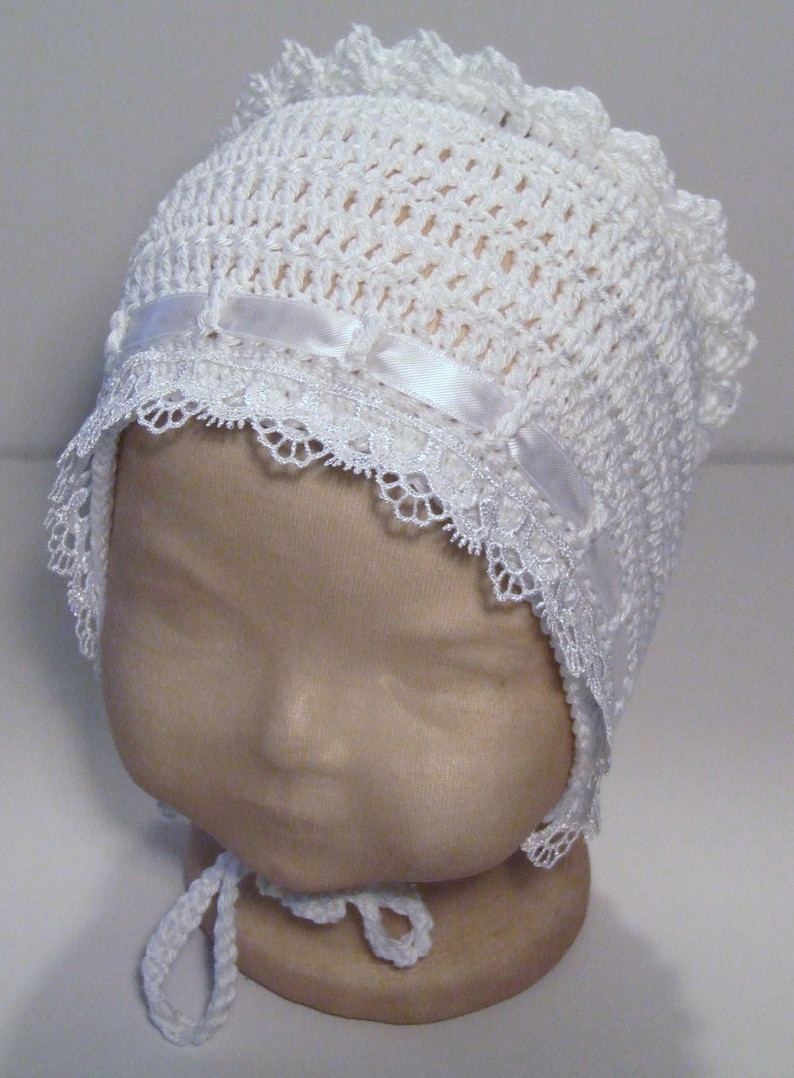 May include: White crocheted baby bonnet with a lace trim and a satin ribbon.
