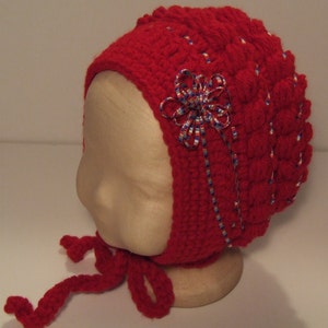 May include: Red crocheted baby bonnet with a white and blue ribbon bow. The bonnet has a tie that can be tied under the chin.