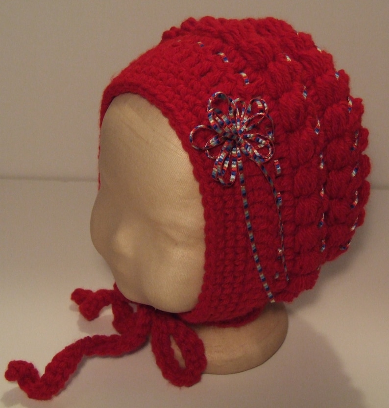 May include: Red crocheted baby bonnet with a white and blue ribbon bow. The bonnet has a tie that can be tied under the chin.