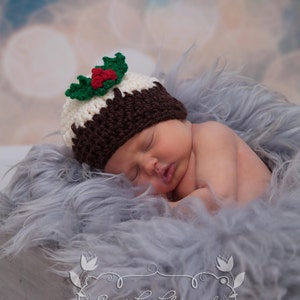 May include: A newborn baby wearing a brown and white crocheted Christmas pudding hat with green holly on top, sleeping on a gray fluffy blanket.