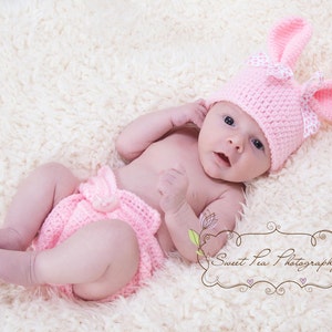 May include: A baby wearing a pink crocheted bunny hat and matching diaper cover. The baby is lying on a white fluffy blanket.