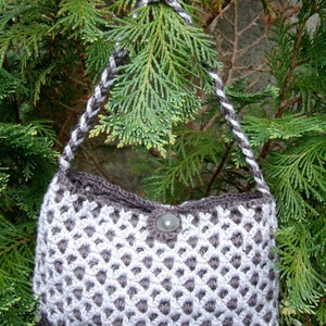 May include: A small, gray and white crocheted handbag with a single button closure. The bag is hanging from a tree branch.