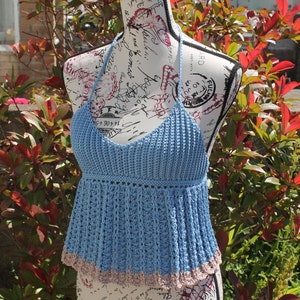 May include: A light blue halter top made of crochet yarn. The top has a fitted bodice and a flared peplum with a contrasting tan trim. The garment is displayed on a mannequin outdoors, surrounded by greenery.