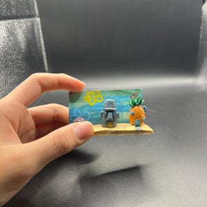 May include: A miniature diorama featuring a painted blue background with yellow flowers, a gray stone-like head sculpture, and a small orange pineapple-shaped house, all mounted on a flat, light-colored rectangular base.