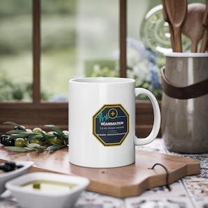 May include: White ceramic mug with a large handle. The mug features a design with the word "RÉANIMATION" in a blue and yellow octagon, with a plus sign and a heartbeat line. The text "Là où chaque seconde compte" is also on the mug.