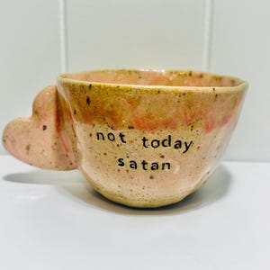 May include: A handmade ceramic mug with a speckled beige and pink glaze, featuring a heart-shaped handle and the text "not today satan" stamped in black lowercase letters on the front. The mug has an irregular, organic shape and texture.