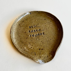 May include: A round, speckled, olive-green ceramic spoon rest with a glossy finish. The centre of the spoon rest features the stamped, lowercase black text that reads: stir... taste... repeat. The item has a slightly raised edge and a small notch.