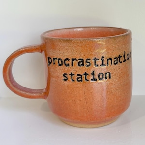 May include: A ceramic mug with a speckled orange glaze and a rounded handle. The text "procrastination station" is printed in black, lowercase, sans-serif letters on the front of the mug. The interior of the mug features a lighter orange tone.