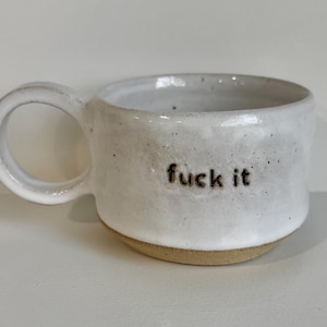 May include: A white ceramic mug with a speckled finish and a round handle. The mug features the text "fuck it" stamped in dark brown lowercase letters on the front. The base of the mug is unglazed, revealing a natural tan clay colour.