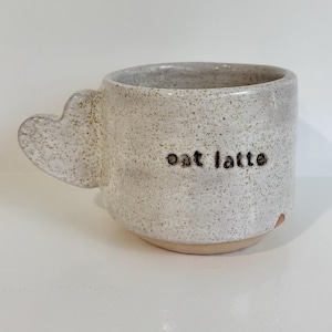 May include: A speckled off-white ceramic mug with a heart-shaped handle. The exterior features the text "oat latte" stamped in dark brown lowercase letters. The base of the mug is unglazed, revealing a light tan clay colour at the bottom edge.