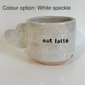 May include: A white speckled ceramic mug with a heart-shaped handle. The text "oat latte" is stamped in black on the front. The base of the mug is unglazed tan clay. The text "Colour option: White speckle" appears at the top of the image.