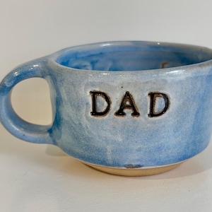 May include: A light blue ceramic mug with a glossy finish and a single handle. The word DAD is embossed in dark, capitalised letters on the front of the mug. The base of the mug is unglazed, showing a natural tan clay colour, while the interior is blue.