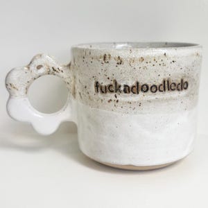 May include: A ceramic mug with a speckled beige upper section and a solid white lower section. The handle is shaped like a flower with rounded petals. The text "fuckadoodledo" is embossed in dark brown letters on the upper side of the mug.