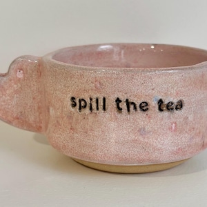 May include: A pink ceramic mug with a speckled texture and a heart-shaped handle. The text "spill the tea" is stamped in black lowercase letters on the front of the mug. The base of the mug is unglazed, showing a natural tan clay colour.