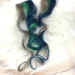 May include: Two long hair extensions featuring a blend of colors including shades of deep forest green, vibrant emerald green, dark navy blue, and light ash brown. The strands are styled in loose, flowing curls that cascade downward.