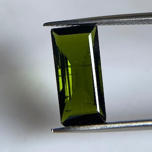 May include: A rectangular, faceted olive green gemstone held by silver-toned metal tweezers. The stone features a clear, transparent structure with visible internal facets and a deep, saturated green hue that transitions to darker tones at the edges.