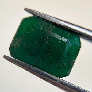 May include: A faceted, rectangular emerald gemstone held by silver tweezers. The deep green gem has visible inclusions and a polished surface, showcasing its natural beauty. The background is a plain, off-white color.