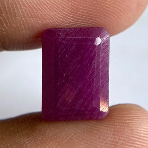 May include: A rectangular, faceted gemstone with a deep magenta-red color. The stone features a clear, polished surface with visible internal linear inclusions and sharp, beveled edges, showcasing a classic emerald-cut style geometry.